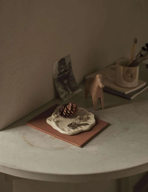 Still life of a pine cone, keys, and a wooden horse figurine on a table with a photo and a mug in the background.