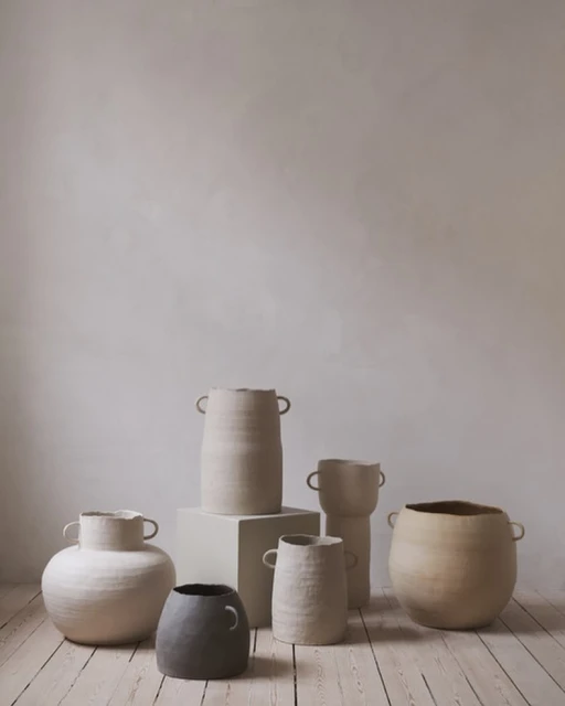 Group of handcrafted clay vases and pots in neutral tones arranged on a weathered wooden floor against a textured wall backdrop.