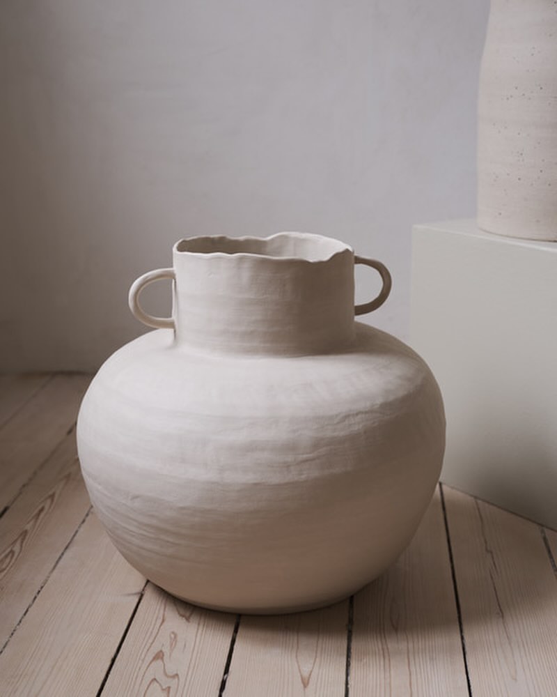 Close-up of a handmade, textured, round, ceramic vase with small handles, sitting on a light wood floor against a textured neutral wall.
