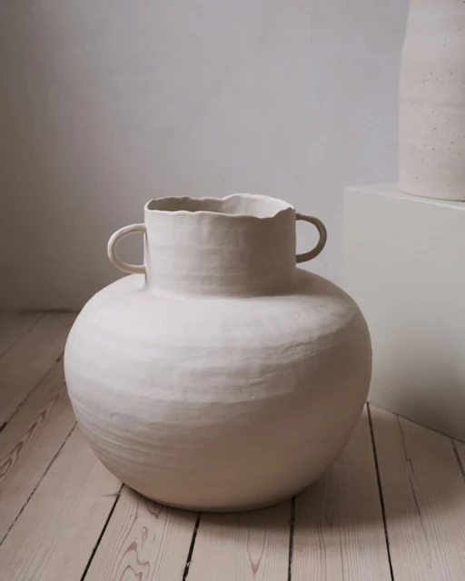 Close-up of a handmade, textured, round, ceramic vase with small handles, sitting on a light wood floor against a textured neutral wall.