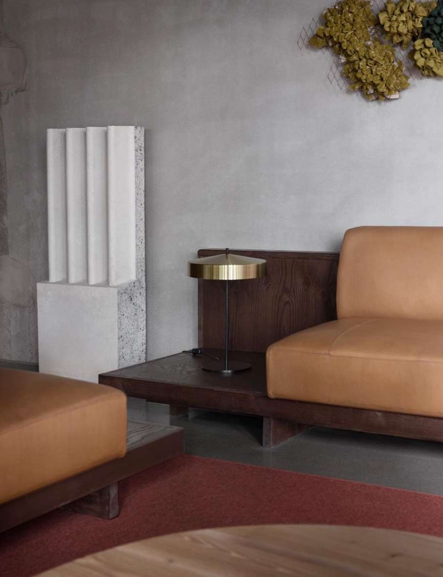 Living room interior featuring a tan leather sofa, a gold lamp, and a decorative stone column against a grey wall with floral decor.