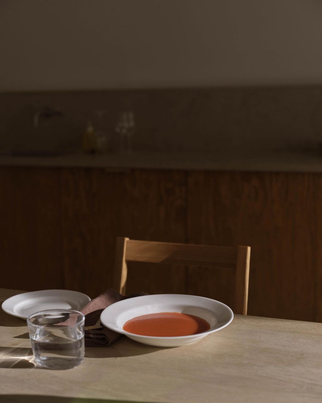 Still life photograph of a dish of tomato soup, a glass of water, and white plates set on a wooden table in a warm, soft light.