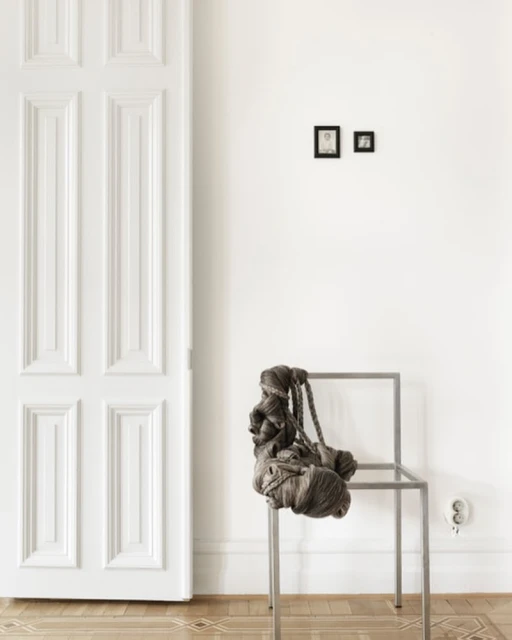 Minimalist interior with white walls, wooden floor, and a modern silver chair draped with a taupe knotted rope decoration or bag.