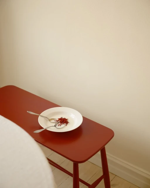Still life of a white plate with red sprinkles and two silver spoons on a red bench against a cream-colored wall indoors.