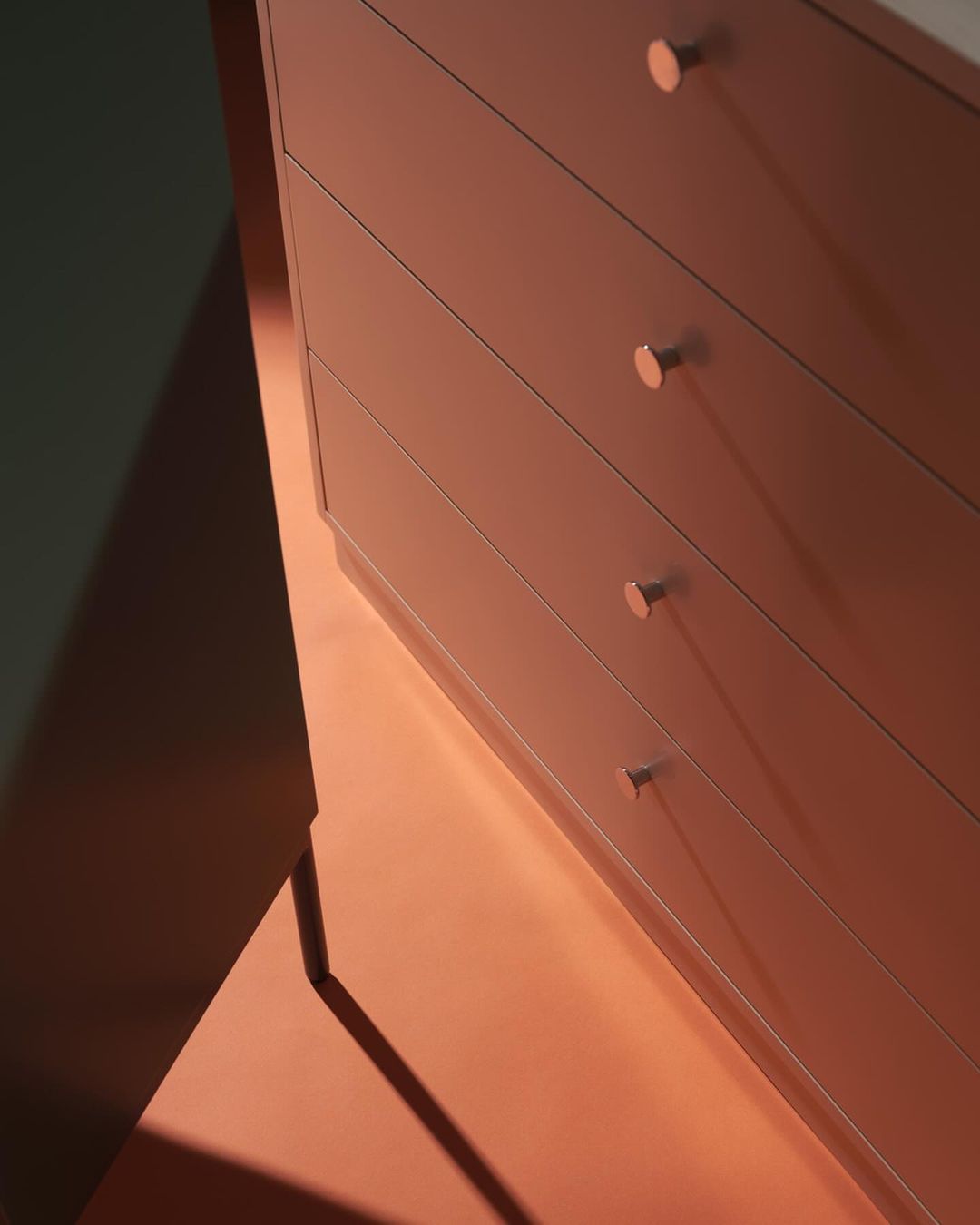 Close-up of a modern light brown dresser with four drawers and round copper handles, casting a long shadow on a coral-colored floor.
