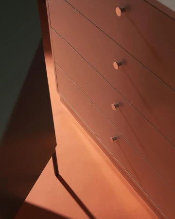 Close-up of a modern light brown dresser with four drawers and round copper handles, casting a long shadow on a coral-colored floor.