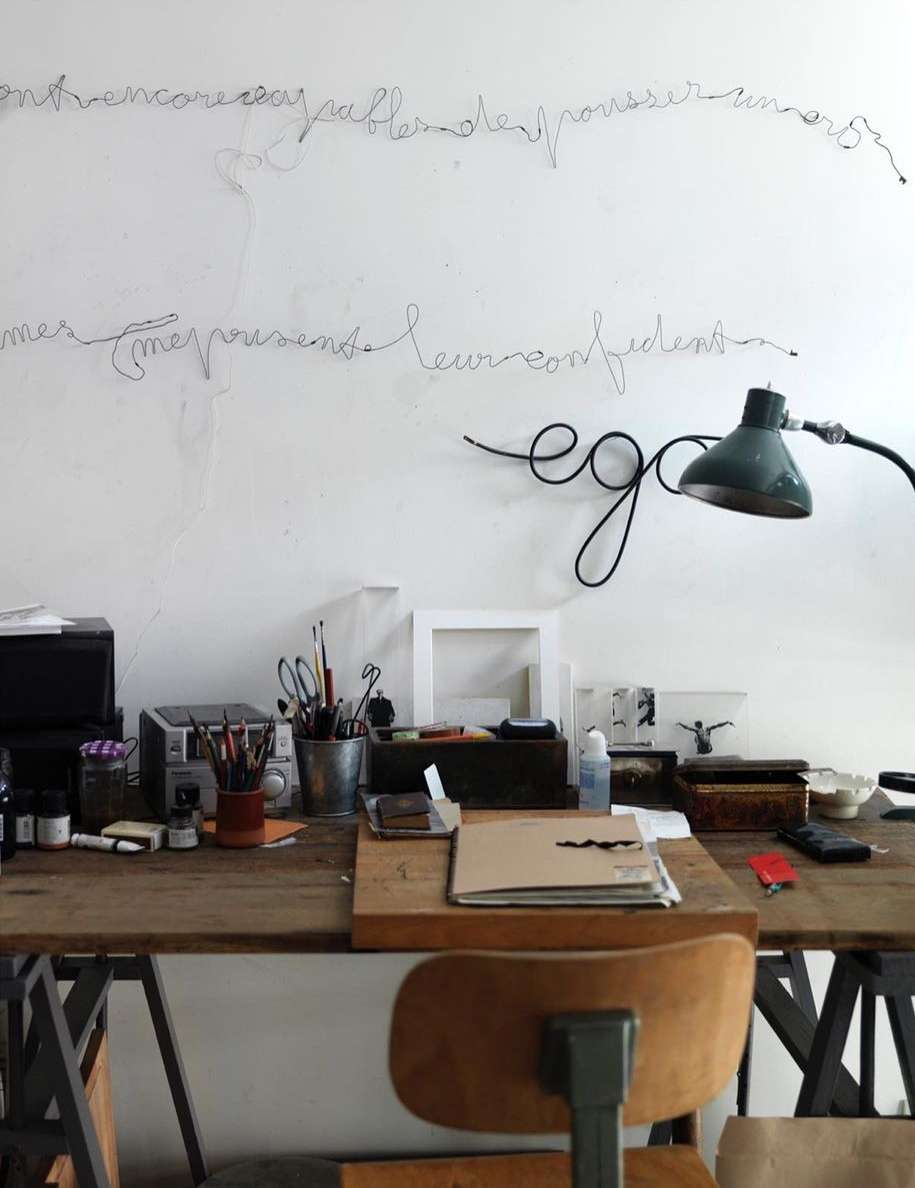 Work desk featuring art supplies, a lamp, and wire art hanging on the white wall, set in an indoor artist studio environment.