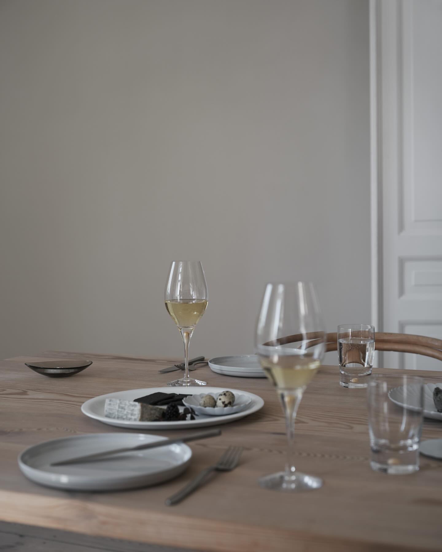 A wood table with a white plates, glasses of white wine, forks, knives, and light gray walls and white trim create a minimalist aesthetic.