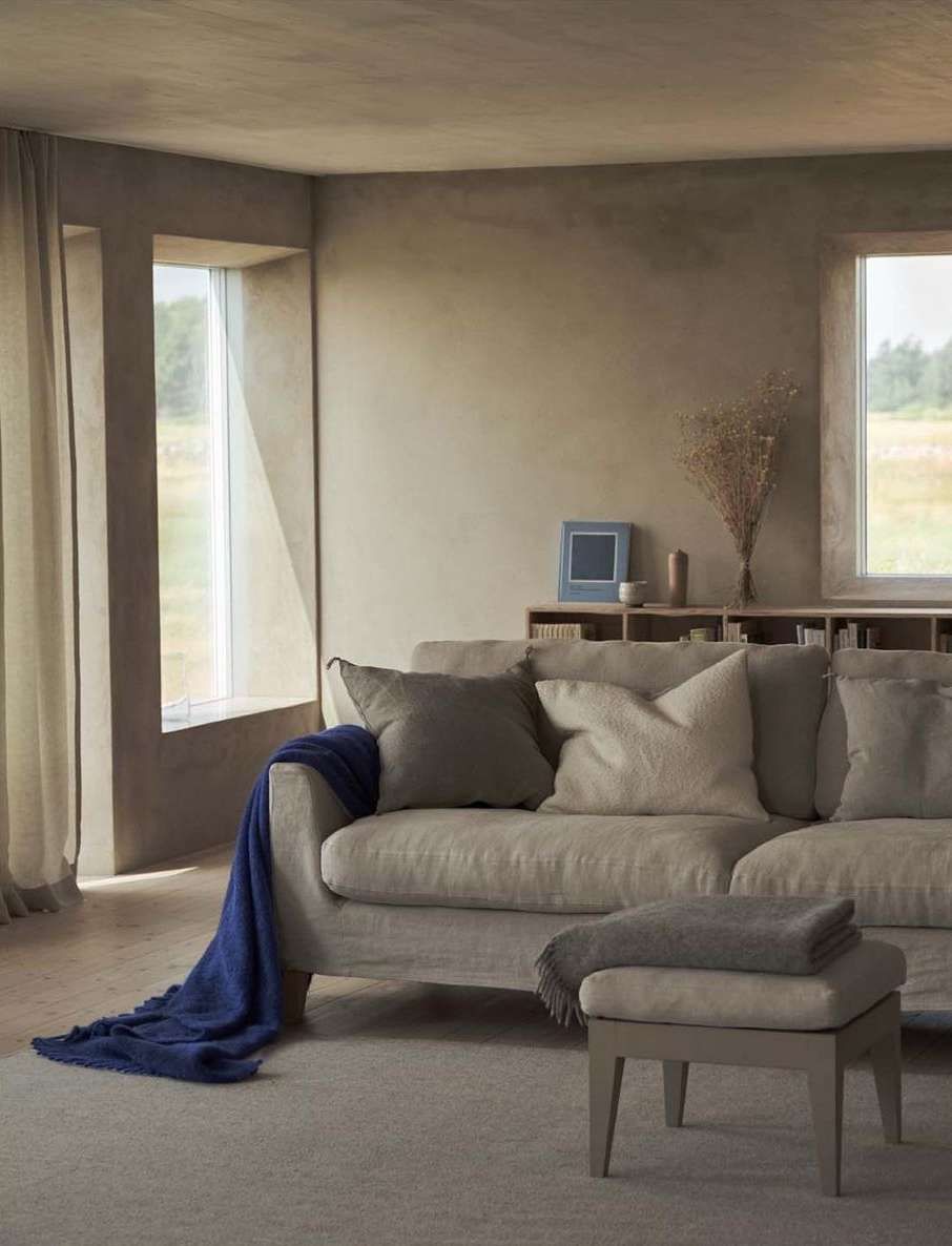 Living room interior featuring a beige sofa with throw pillows, a dark blue throw blanket, and a light-colored ottoman in a neutral space.