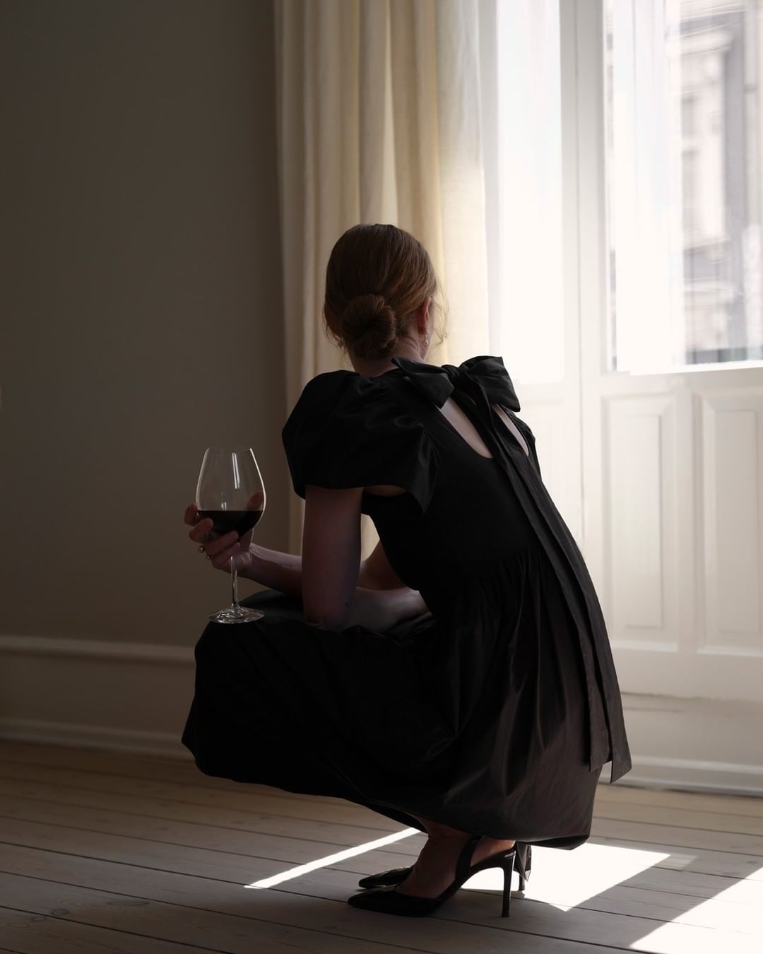 Woman in black dress crouching near a window holding a wine glass by photographer Petra Bindel