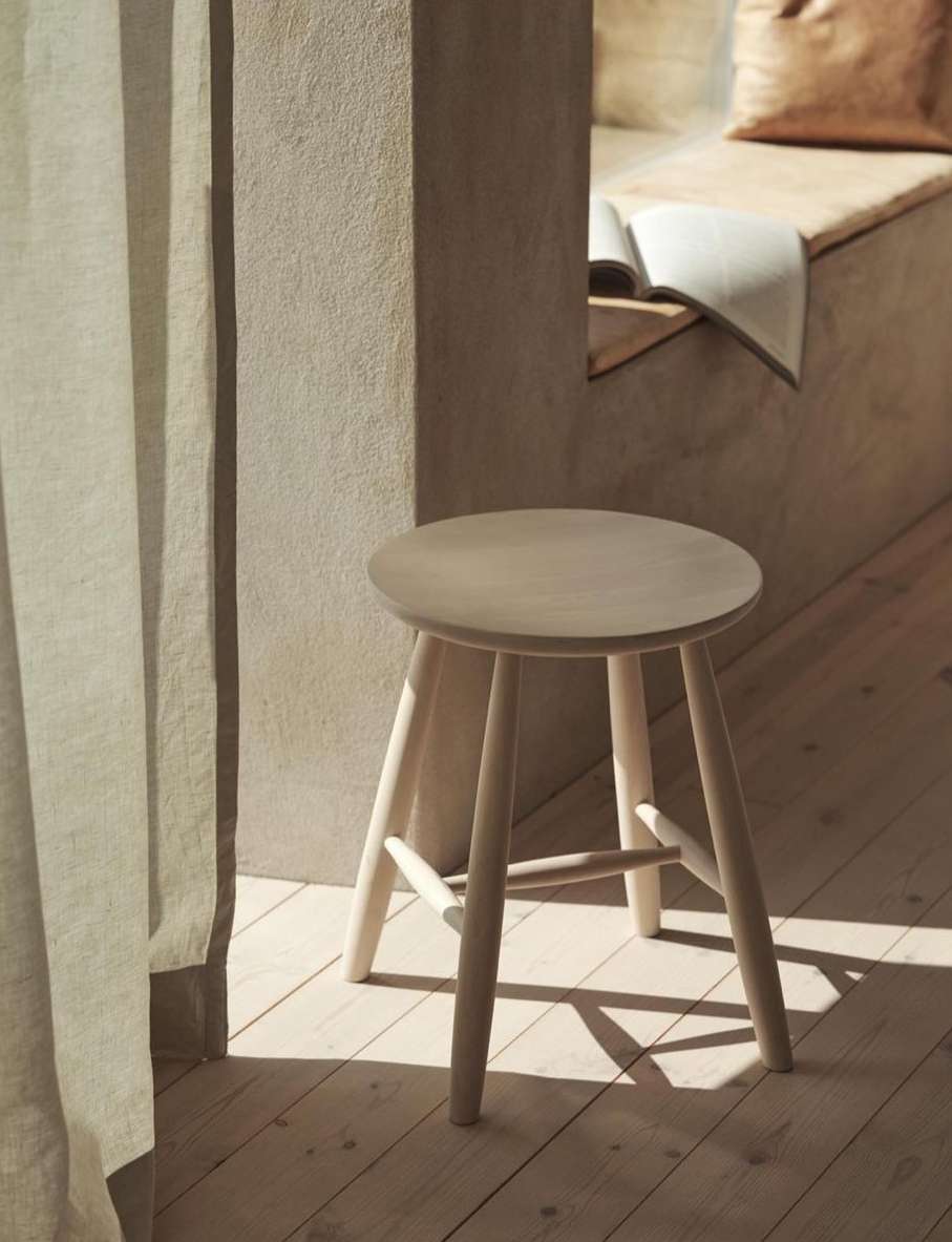 Neutral indoor shot featuring a minimalist wooden stool set on a light-toned wooden floor, complemented by a window seat and linen curtains.