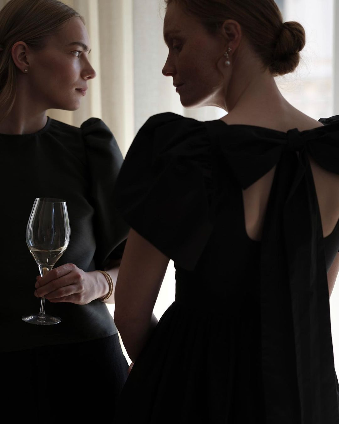 Two fair-skinned adult women pose indoors, one in a black dress with a large bow, the other holding a glass of white wine.