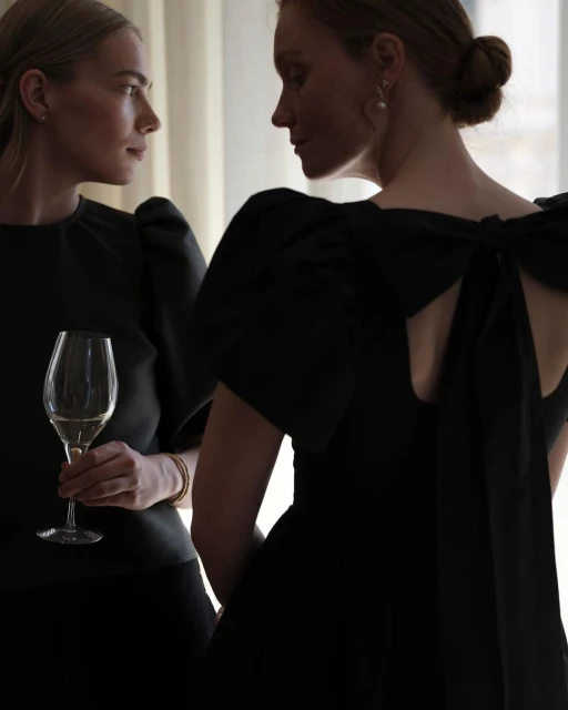 Two fair-skinned adult women pose indoors, one in a black dress with a large bow, the other holding a glass of white wine.