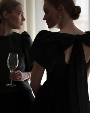 Two fair-skinned adult women pose indoors, one in a black dress with a large bow, the other holding a glass of white wine.