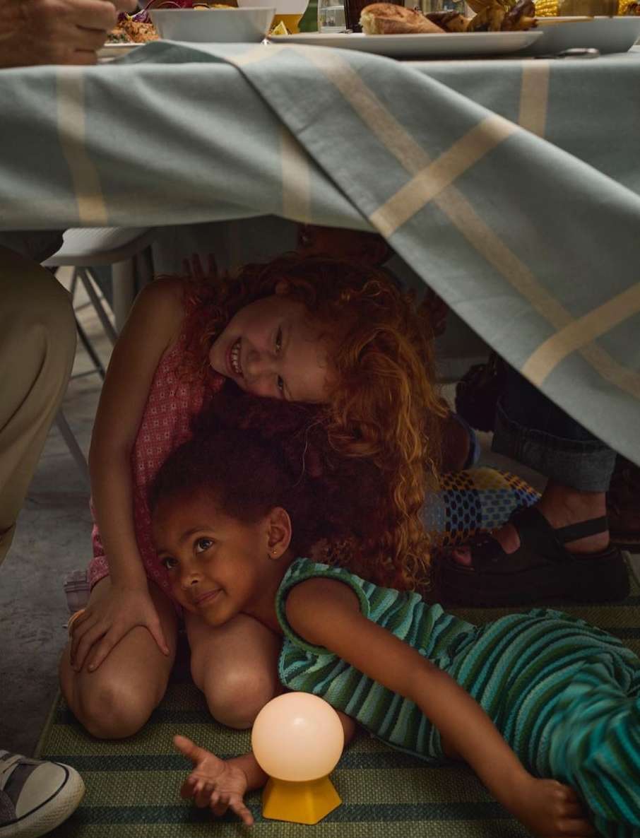 Two young children, one Black and one white, lay under a table covered with a blue and white tablecloth during a meal.