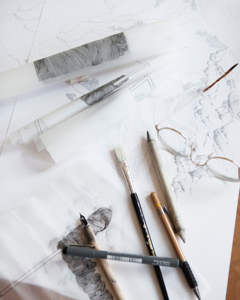 Overhead shot of illustration tools and drawings on a white surface: pencils, pens, brushes, eyeglasses, and rolled-up sketches.