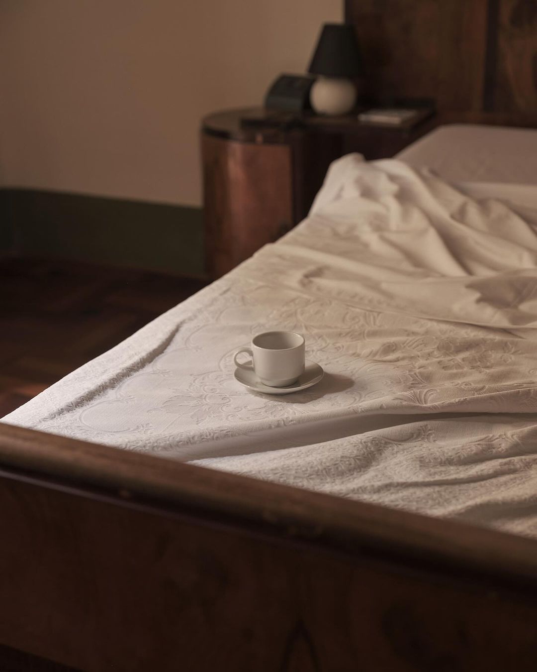 Cozy bedroom scene with a white cup and saucer on a patterned bed, wooden frame, bedside table, and lamp in soft, warm lighting.