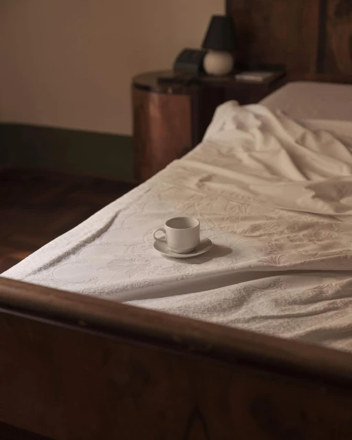 Cozy bedroom scene with a white cup and saucer on a patterned bed, wooden frame, bedside table, and lamp in soft, warm lighting.