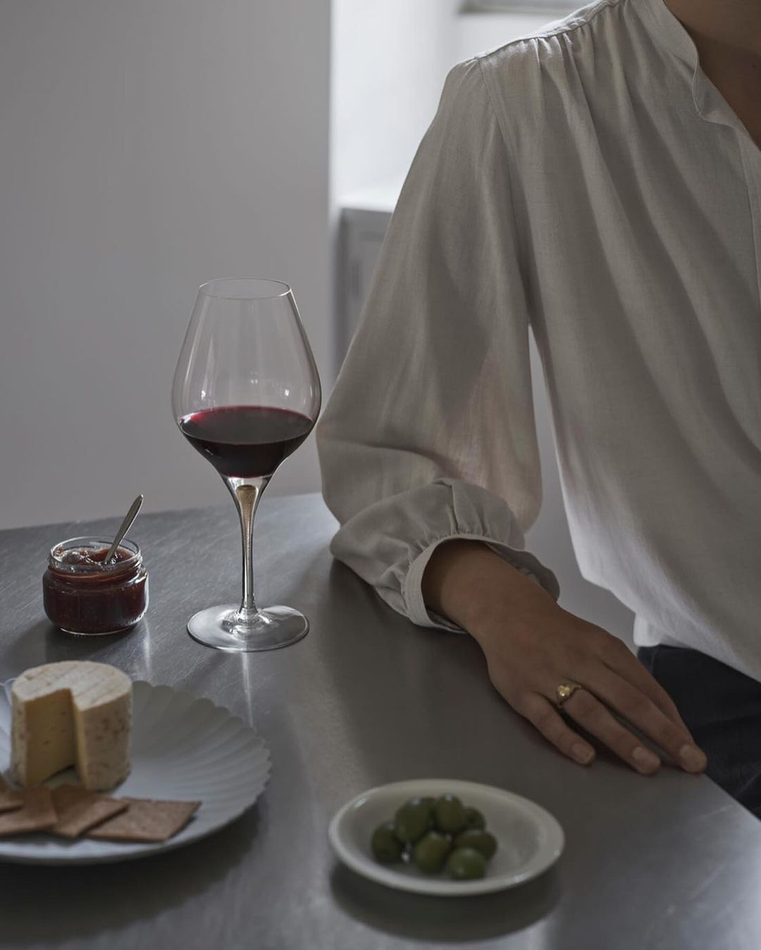 Still life shot of red wine, cheese, crackers, olives, and jam arranged on a counter with a person partially visible next to them.