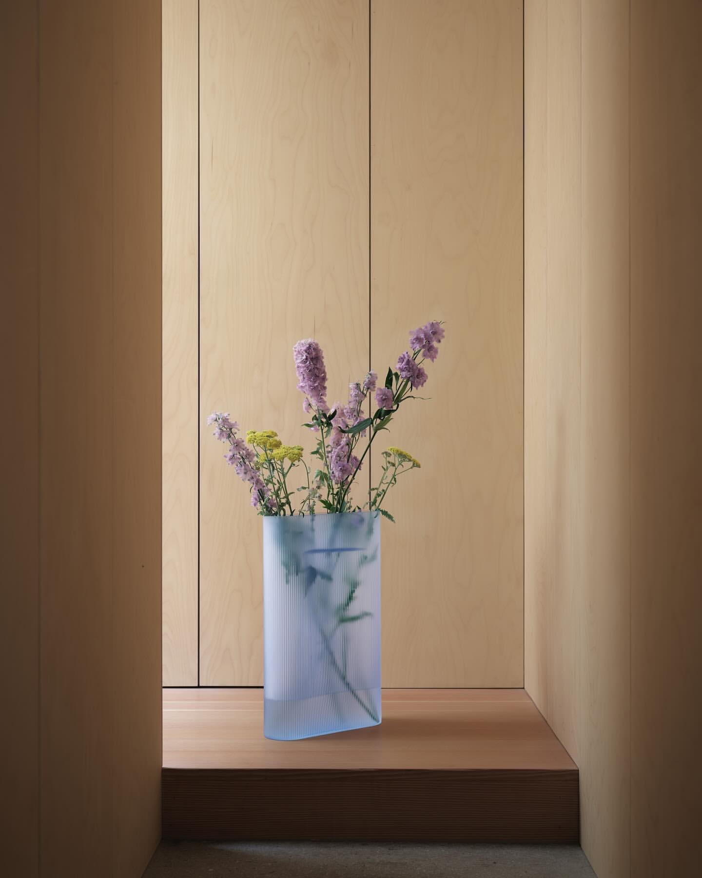 Indoor still life: lilac and yellow flowers in a light blue geometric vase on a wood surface against a light wood panel wall.