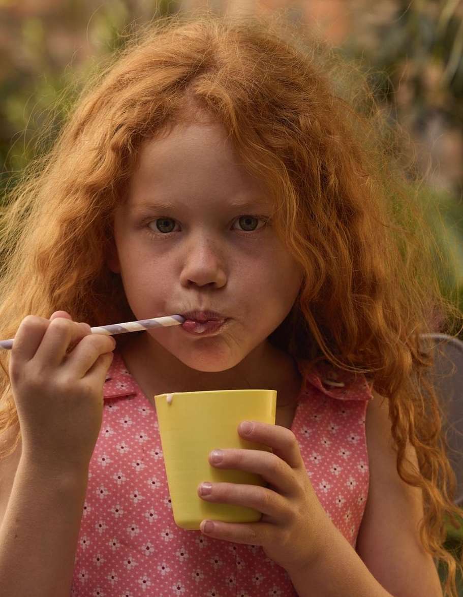 Close-up shot of a young girl with red curly hair eating yogurt from a yellow cup with a straw outdoors in daytime.