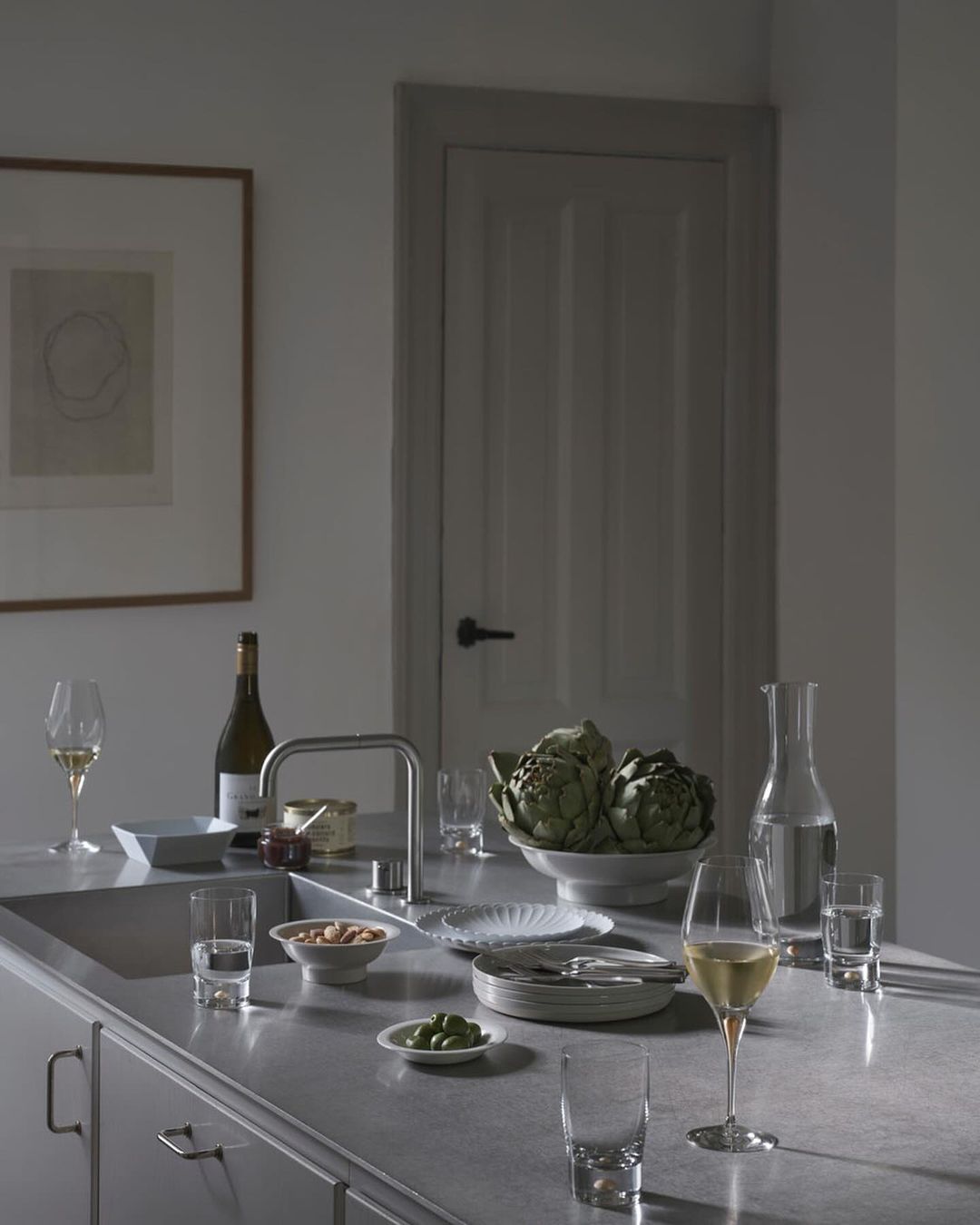 Still life on a kitchen counter features wine, artichokes, nuts, plates, glasses, water, and silverware against a neutral wall and door.