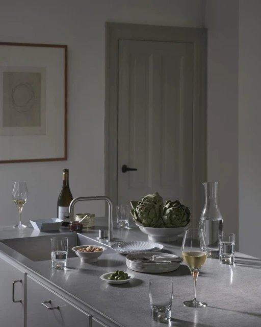Still life on a kitchen counter features wine, artichokes, nuts, plates, glasses, water, and silverware against a neutral wall and door.