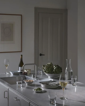 Still life on a kitchen counter features wine, artichokes, nuts, plates, glasses, water, and silverware against a neutral wall and door.