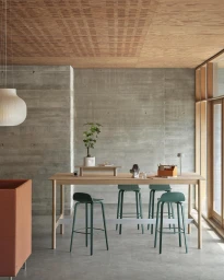 Minimalist interior with a long wooden table, green stools, concrete wall, and wooden panel ceiling photographed by Petra Bindel.