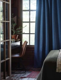 Interior view of a cozy room with a dark wooden desk, leather chair, potted plant, and a blue curtain covering a multi-pane window.