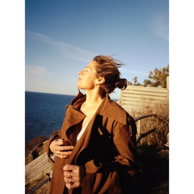 Woman in a brown coat stands with her face to the sun near the ocean under a blue sky, exuding a sense of freedom and serenity.