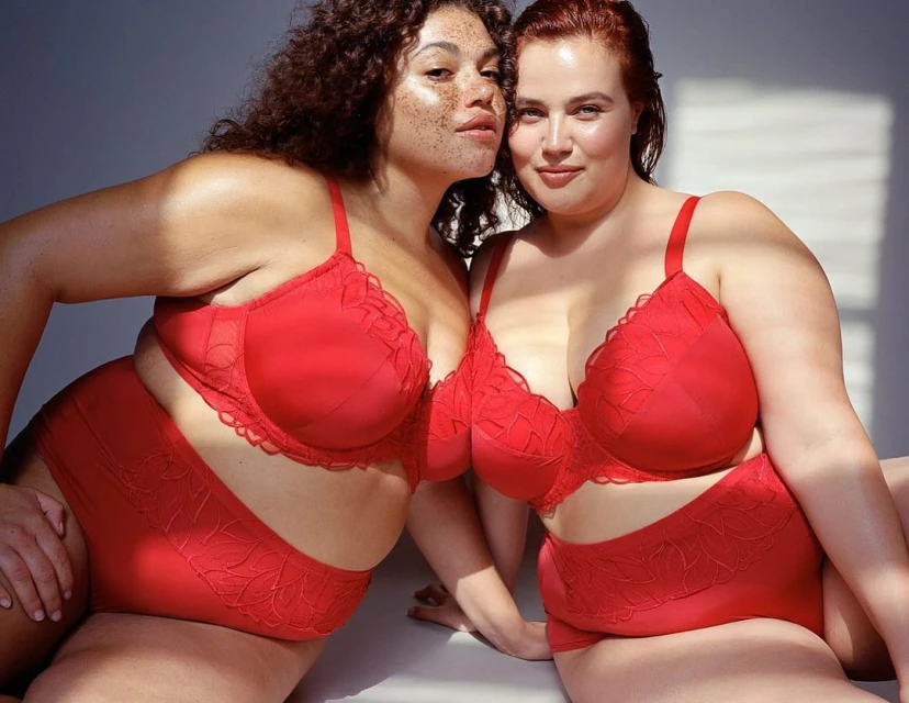 Two plus-size women in matching red lingerie pose in a brightly lit indoor studio with soft shadows, showcasing body positivity and confidence.