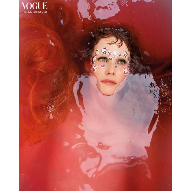 Portrait of a woman partially submerged in reddish liquid, adorned with stickers on her face, set against a background of rippling liquid.