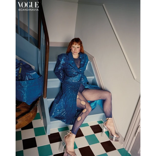 A redhead fashion model poses seated on blue stairs wearing a shimmering blue dress and tights, paired with platform heels, for Vogue.