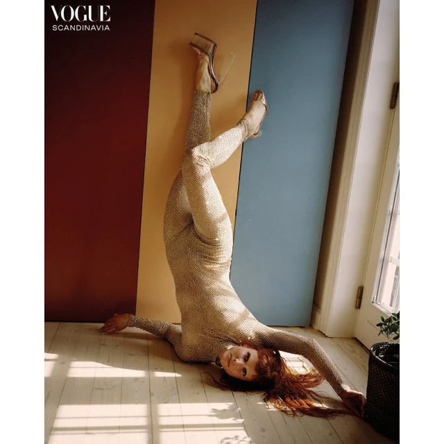 A red-haired fashion model in a gold sequin jumpsuit poses upside down in front of a colorful wall in a studio with natural light.