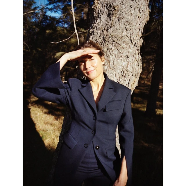 Woman in a navy suit stands by a large tree in a forest, shielding her eyes from the sun with her hand, looking off into the distance.
