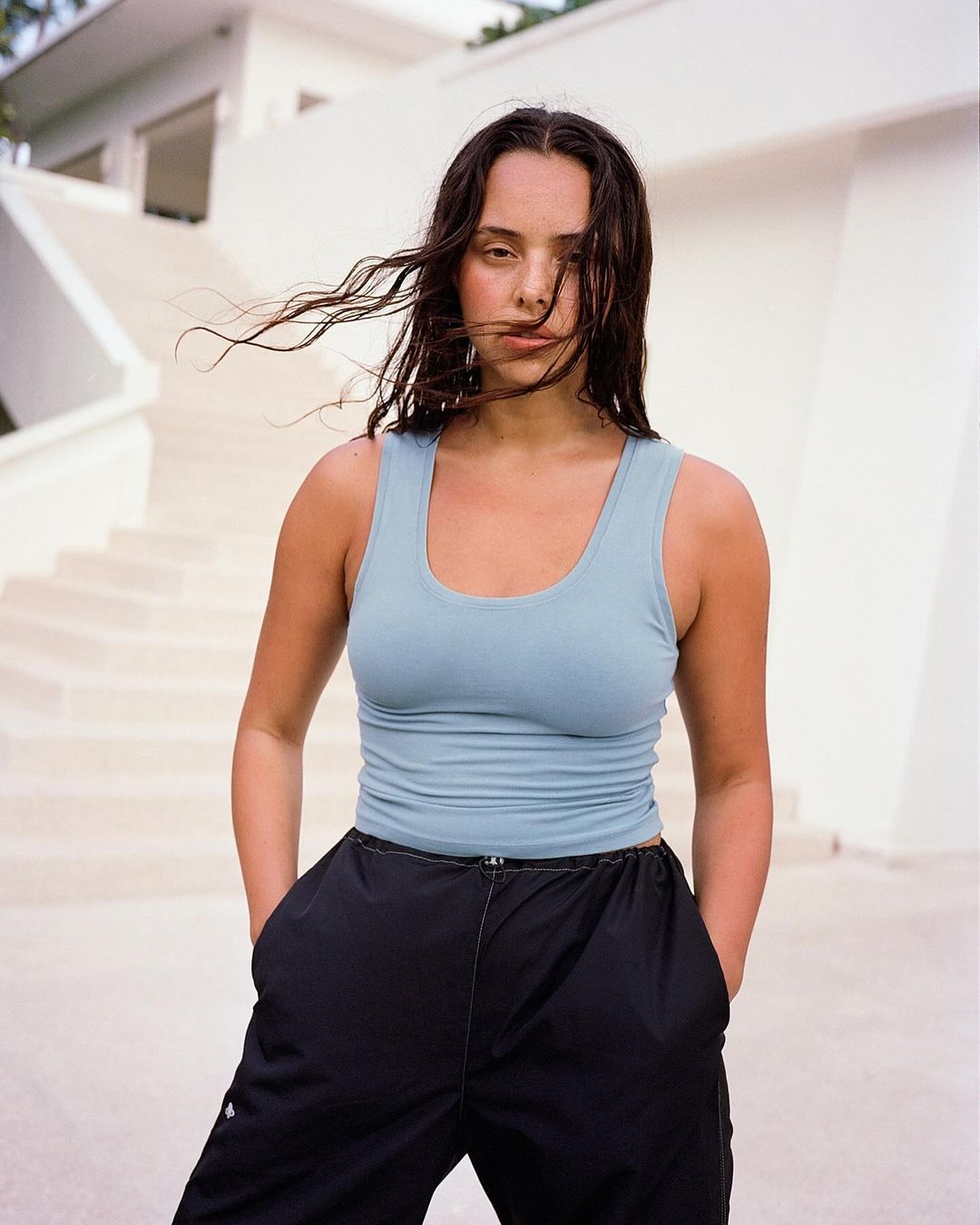 A woman with wind-blown hair is posing outside in a light blue tank top and black pants in front of a modern white building.