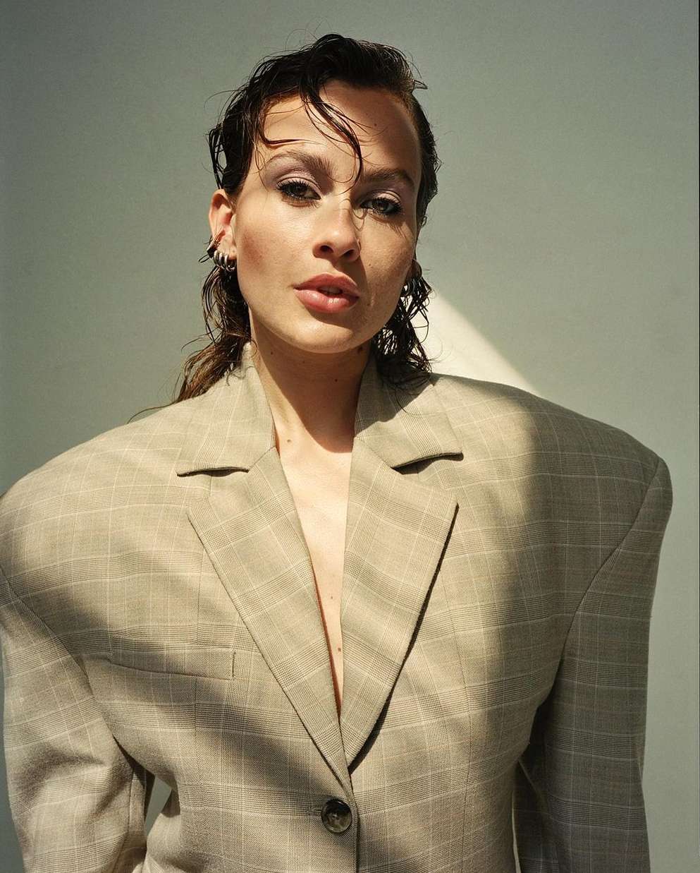 A young woman in an oversized beige plaid blazer poses indoors, with wet-look hair and natural makeup in a medium shot against a neutral wall.