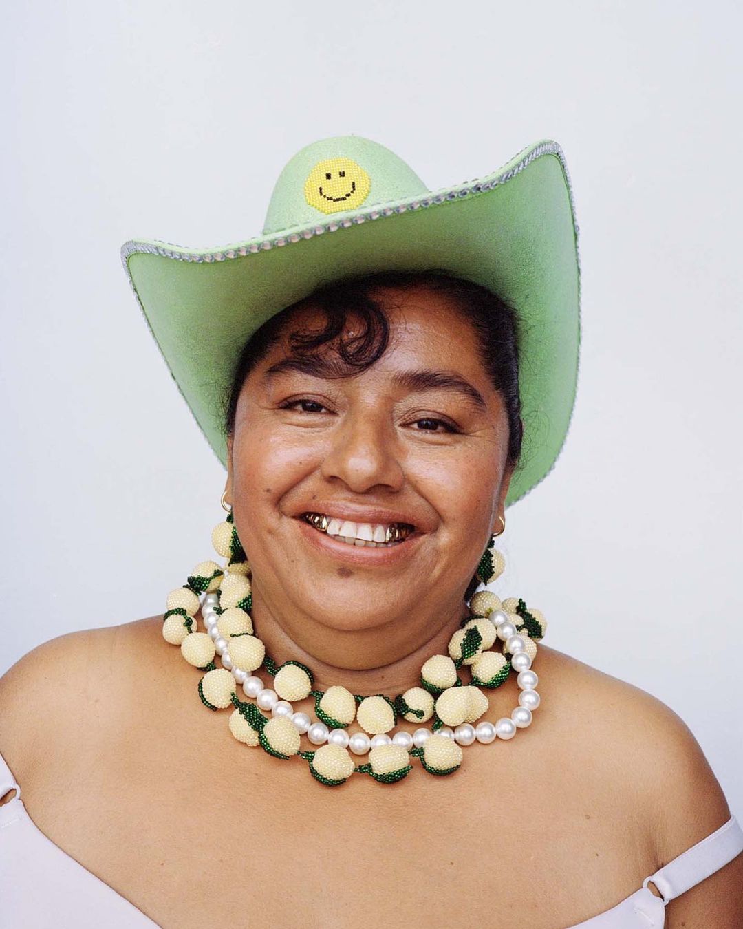 Smiling woman wearing a green cowboy hat and beaded necklace in a studio portrait against a white backdrop. Fashion and portrait photography.