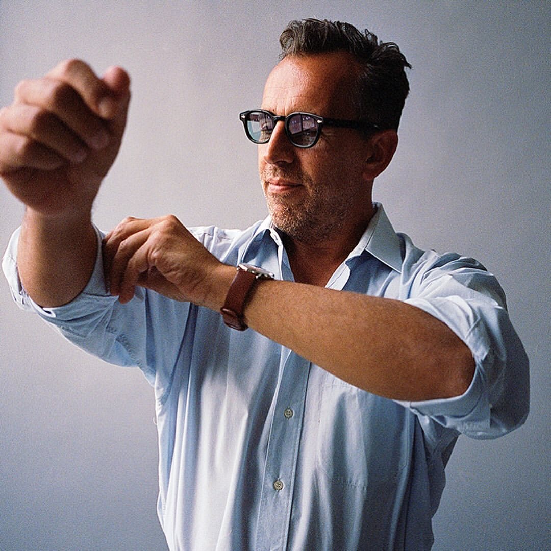 Man wearing round black sunglasses and a light blue button-down shirt rolling up his sleeve against a plain grey backdrop.