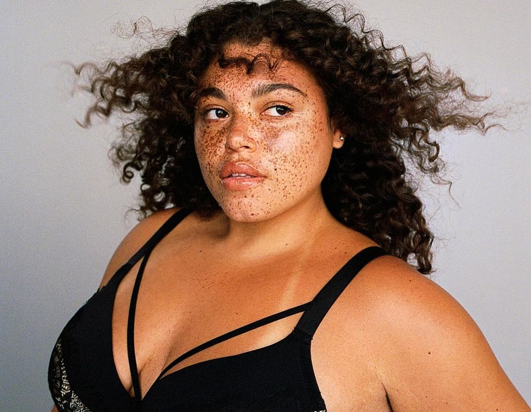 Portrait of a woman with freckles, wearing a black bra, with curly brown hair against a neutral background in a beauty context.