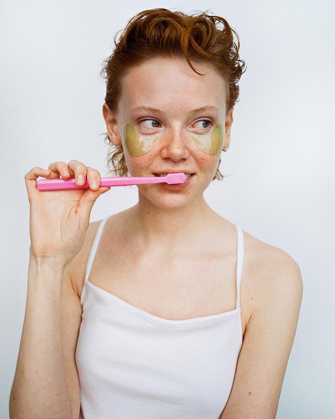 A young red-haired woman with eye masks holds a pink toothbrush, standing in front of a clean white background, beauty and dental care.