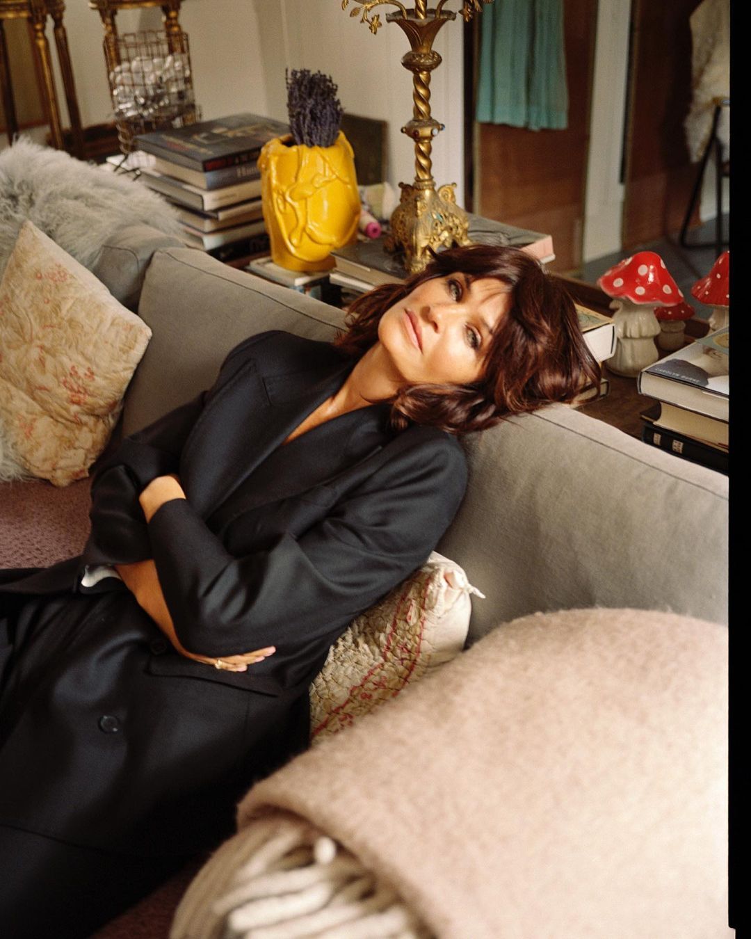 Woman with dark hair lounges on a gray sofa in a black blazer, with a blanket, books, and decorative objects in a home interior.
