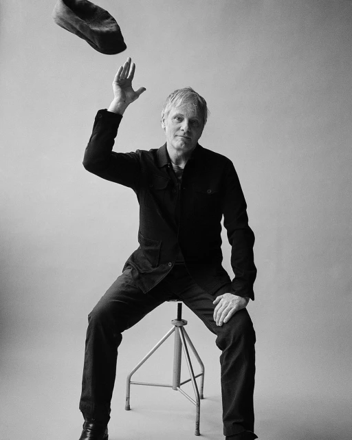 Black and white portrait of an adult male seated on a stool, tossing a hat into the air against a light grey backdrop in a photo studio.