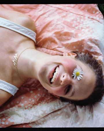 Portrait by Petra Kleis of a smiling woman lying on a pink patterned blanket with a daisy flower placed over her closed eye