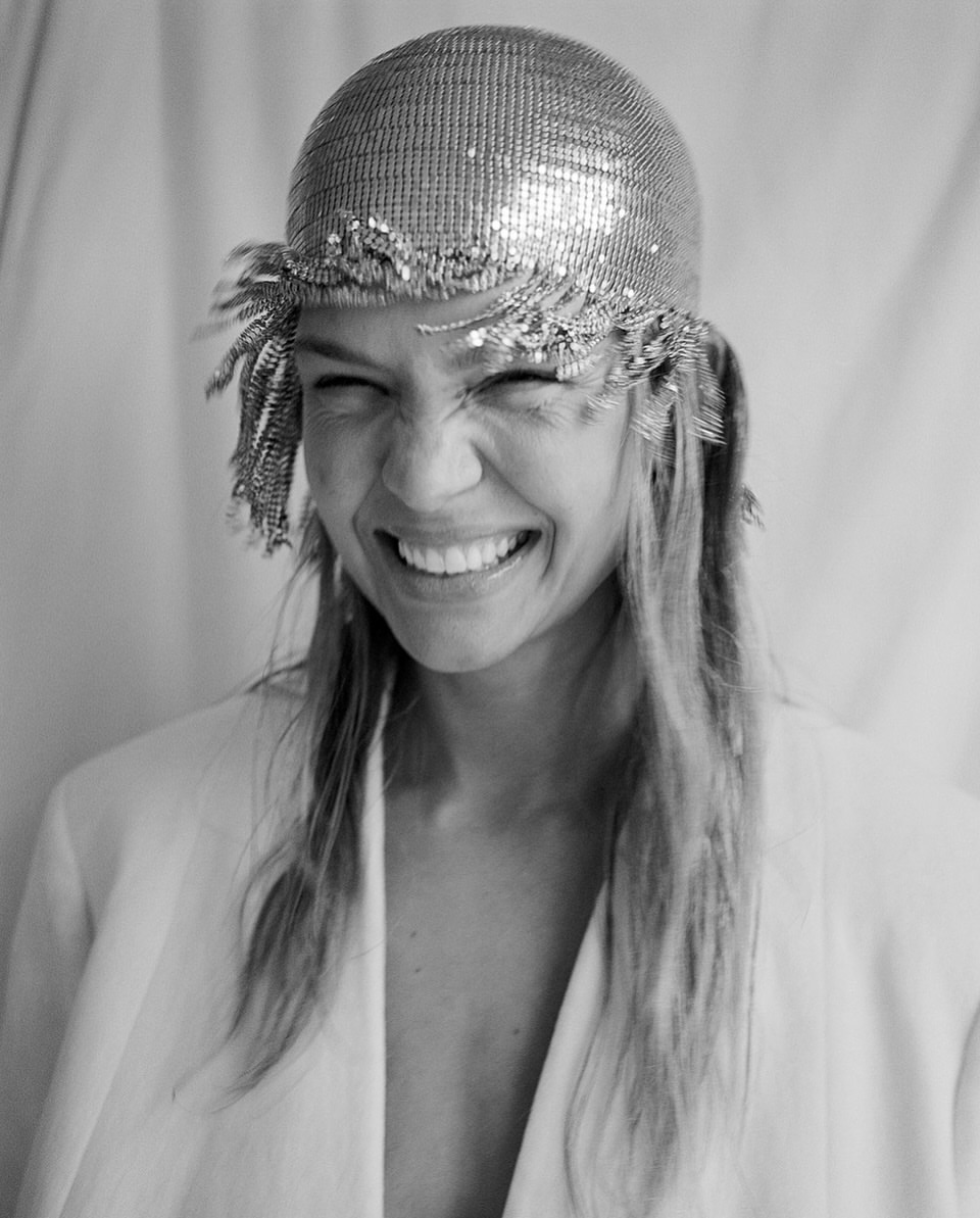 Close-up shot of a smiling adult woman wearing a metallic beaded cap and white jacket against a draped white backdrop, studio lighting.