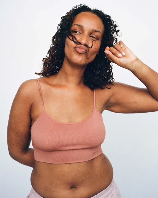 Portrait of a young adult woman with curly hair wearing a crop top and light pink bottoms, posing against a white backdrop.