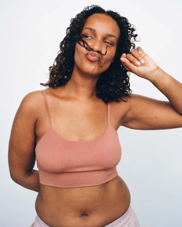 Portrait of a young adult woman with curly hair wearing a crop top and light pink bottoms, posing against a white backdrop.