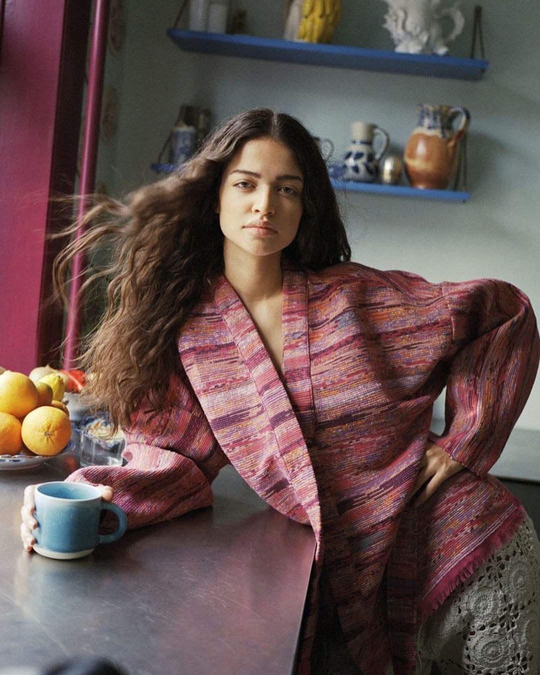 A young woman with long, curly brown hair rests her arms on a kitchen counter while holding a blue mug. The image shows a warm, lifestyle aesthetic.