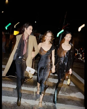 Three models in fashionable attire stroll on a city crosswalk at night, one in a beige coat, another in a black dress, and one in sequins.